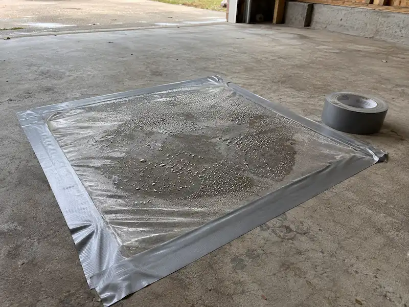 Square of clear plastic taped to a garage floor with water droplets under it and darker concrete beneath