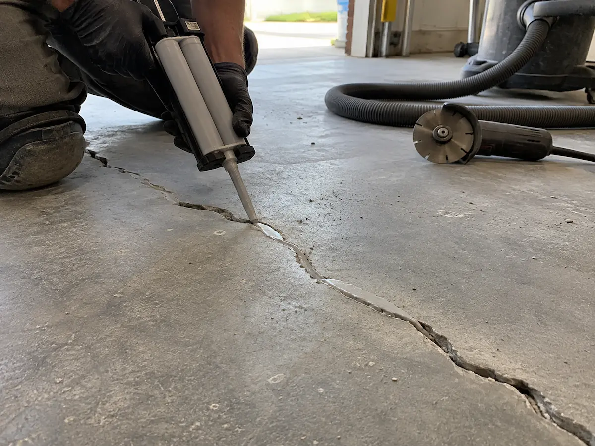 Gloved hands filling a cleaned-out crack in a garage floor with repair material from a dual-cartridge gun