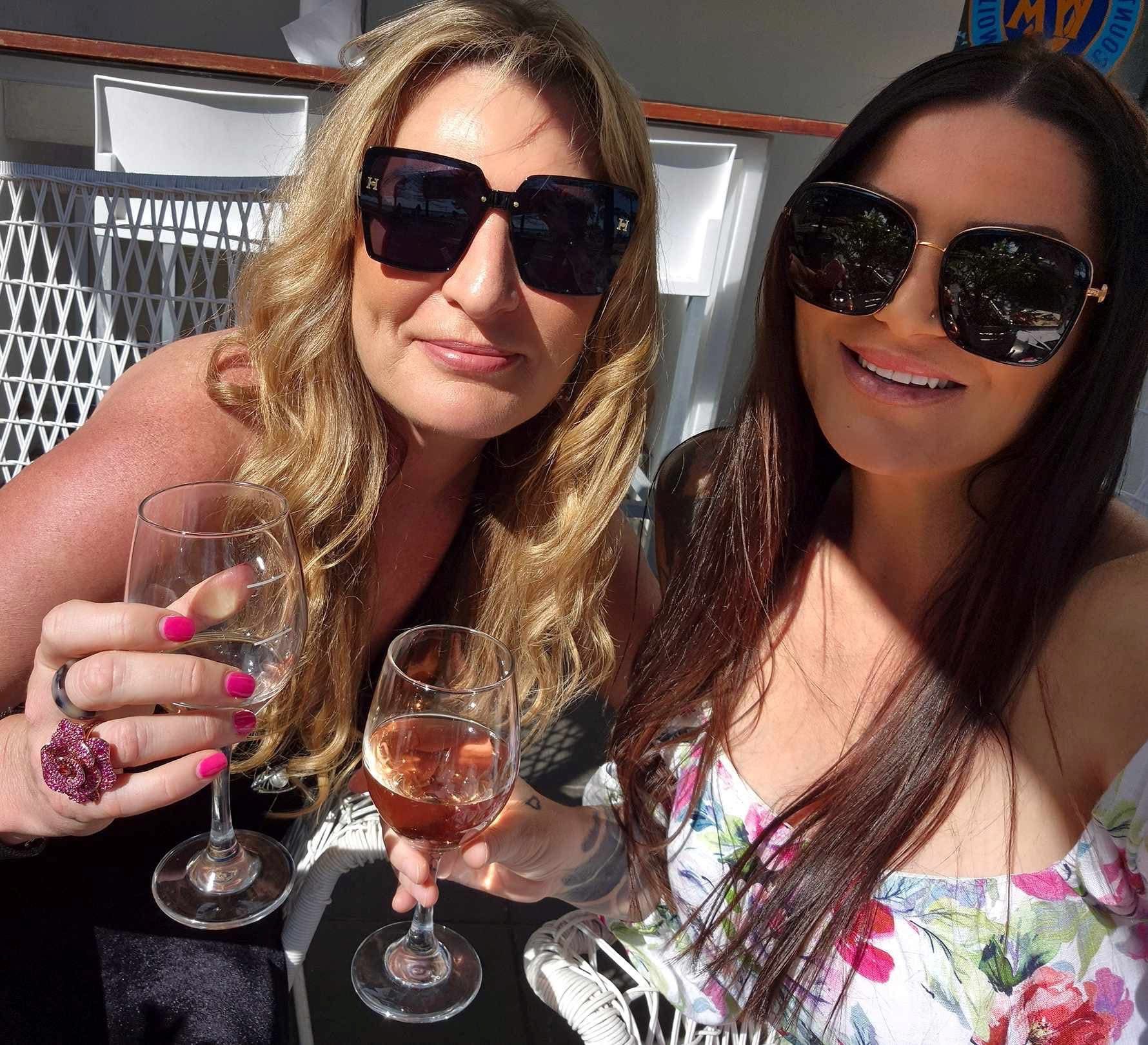 Two friends in sunglasses smiling for a selfie with glasses of wine on a sunny terrace