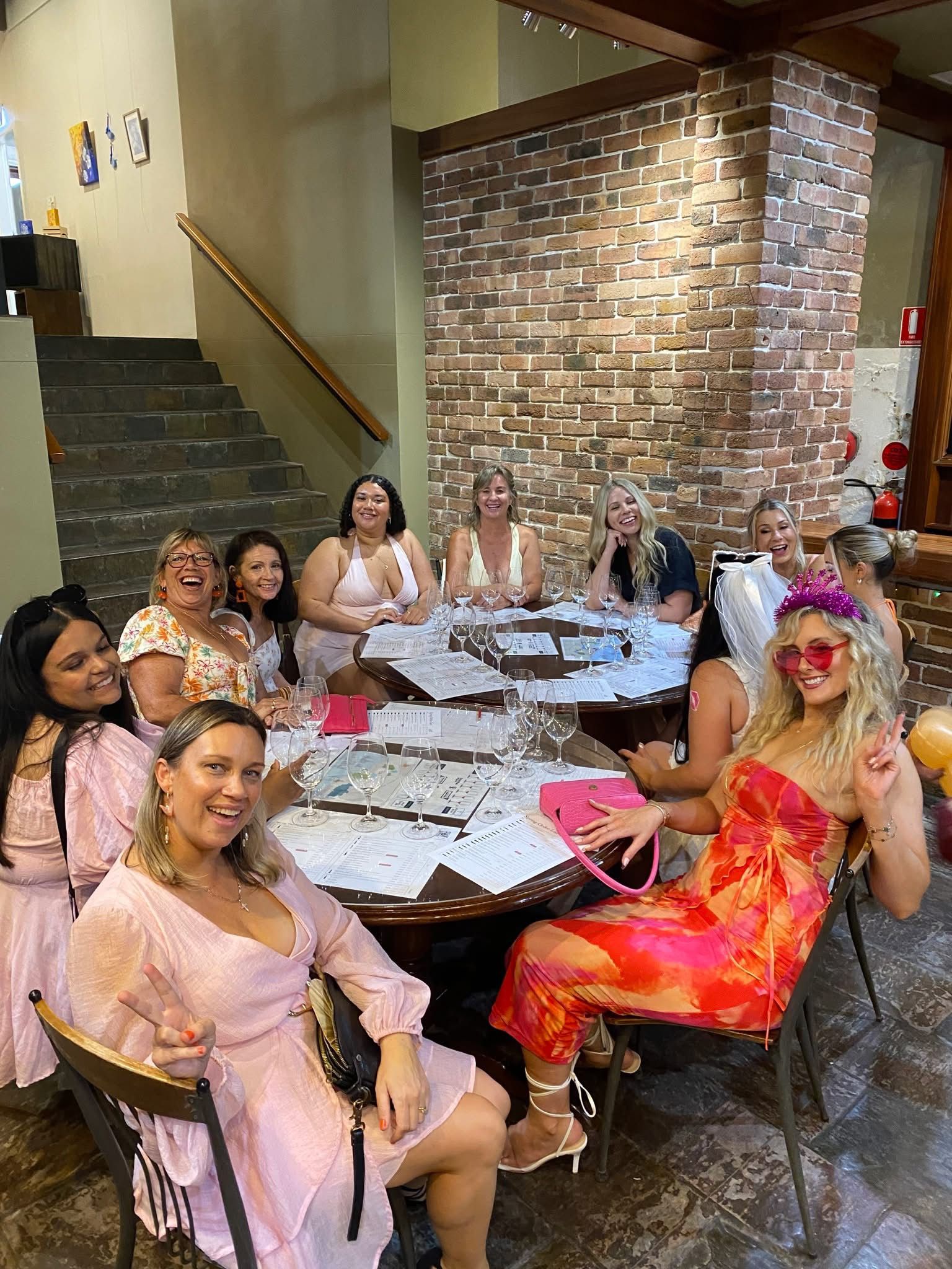 A hens group seated around a tasting table, with the bride to be in pink heart sunglasses and a bright orange dress smiling on the right