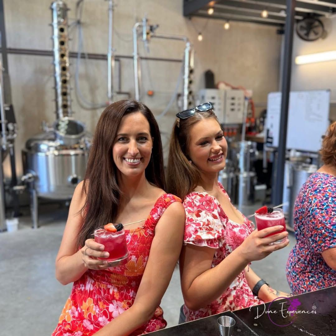 Two friends holding pink cocktails inside a distillery with copper stills behind them