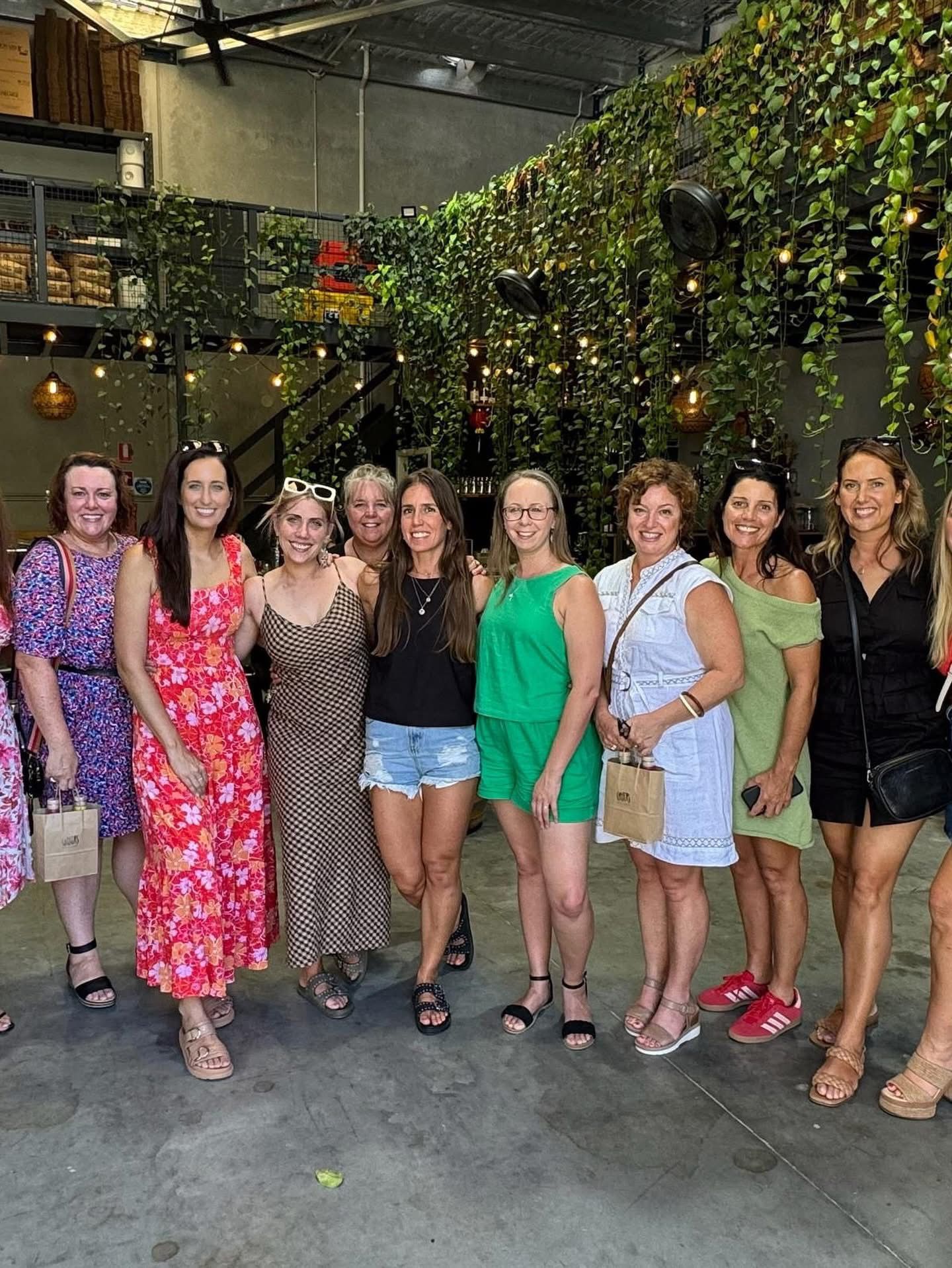 A group of women in summer dresses smiling together in front of a wall of hanging greenery at a venue