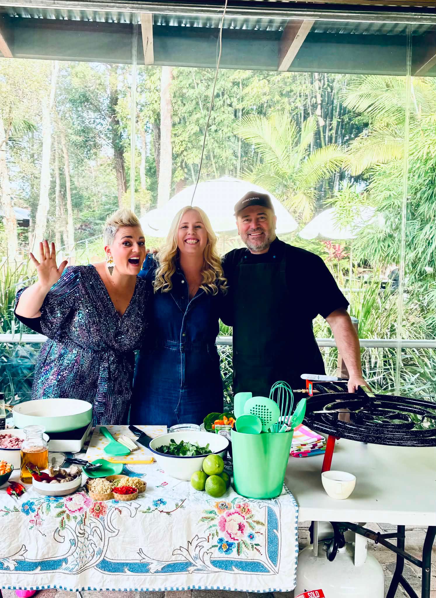 Three hosts smiling behind a table of fresh produce at an outdoor cooking demonstration in a leafy garden