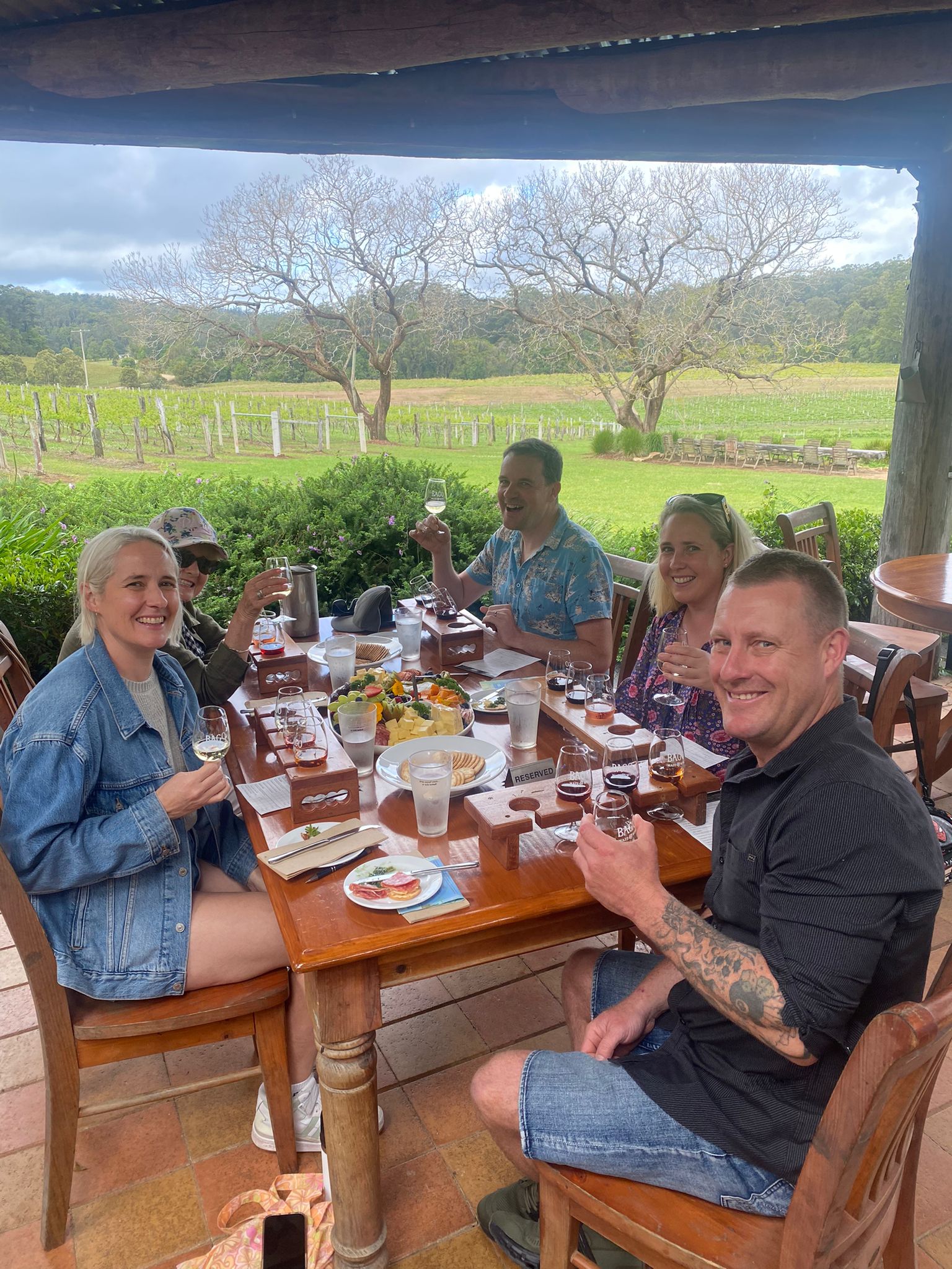 Guests talking and laughing around a long shared table on a covered deck overlooking green paddocks