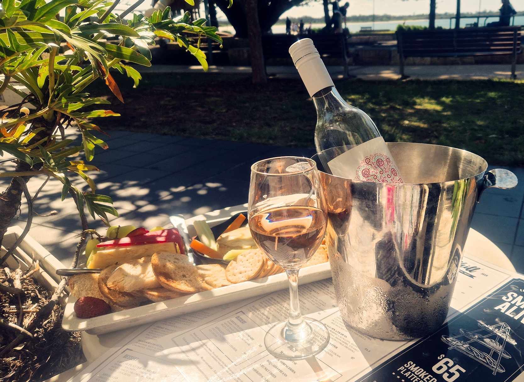 Two glasses of rose beside a chilled bottle and a grazing plate on an outdoor table set for two
