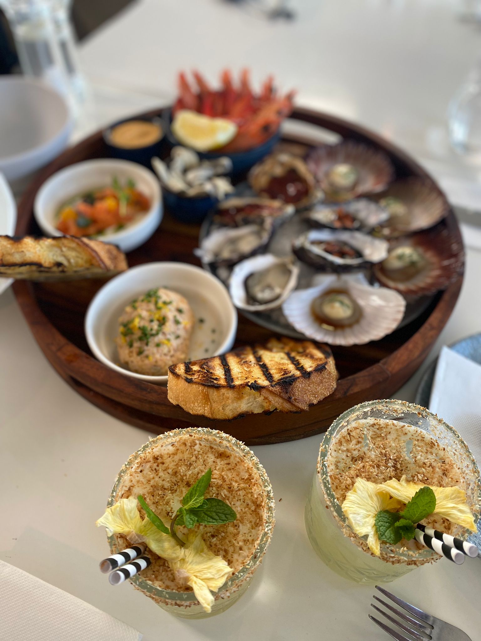Shared plates of oysters, dips and bread with two mocktails on a white table
