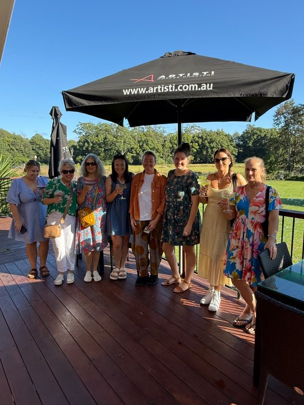 A group of eight women with drinks smiling together on a sunny deck under a large umbrella
