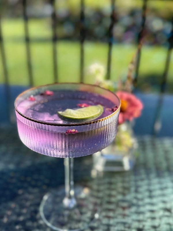 A purple drink in a coupe glass, garnished with lime and flower petals