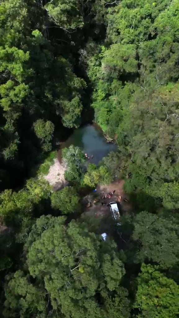 Aerial view over rainforest to a swimming hole, with a white minibus parked nearby