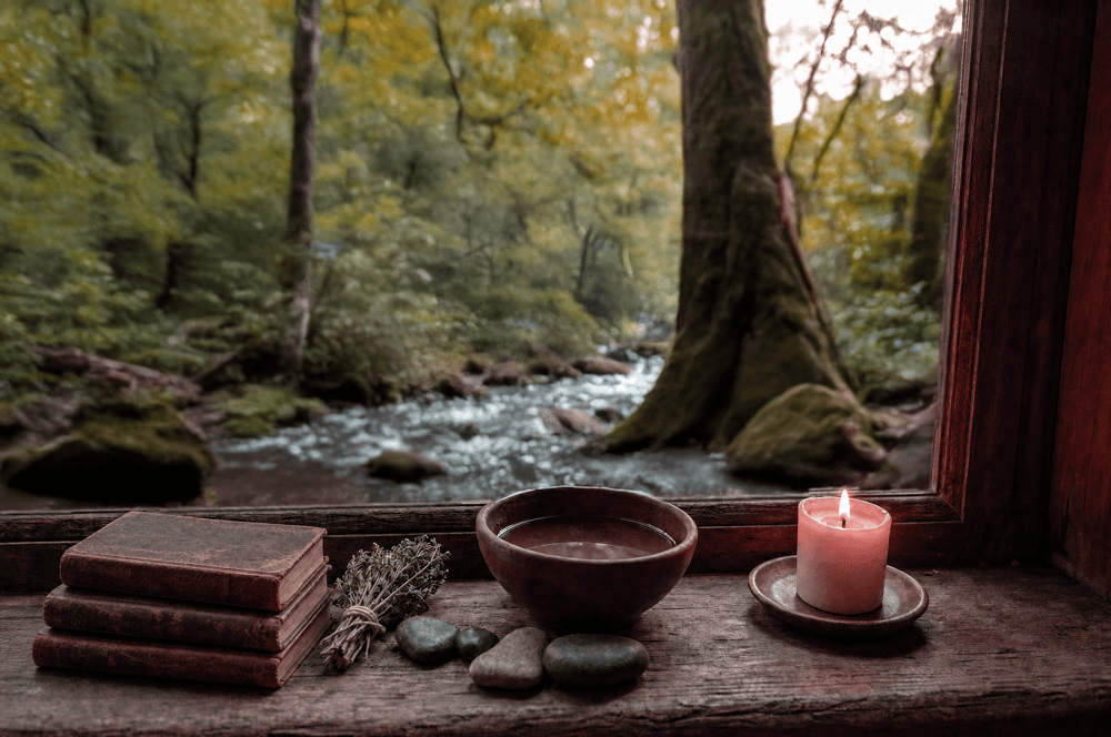 A lit candle, three old books, and a clay bowl on a wooden windowsill, open to the cloud forest and a stream