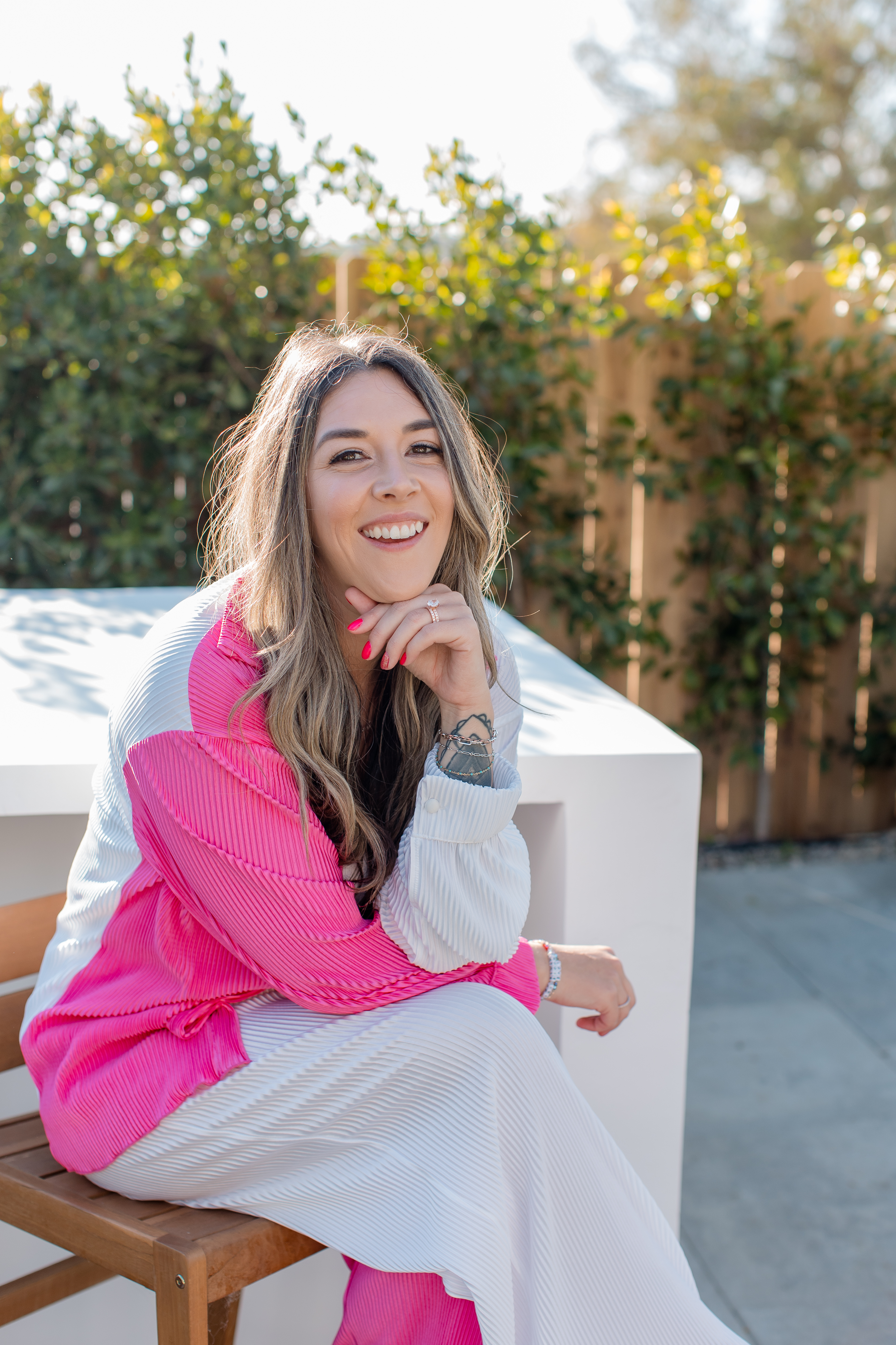 Shelby St. Clair smiling outdoors in a bright pink and white outfit, sitting at a modern white table — representing confidence, joy, and aligned entrepreneurship.