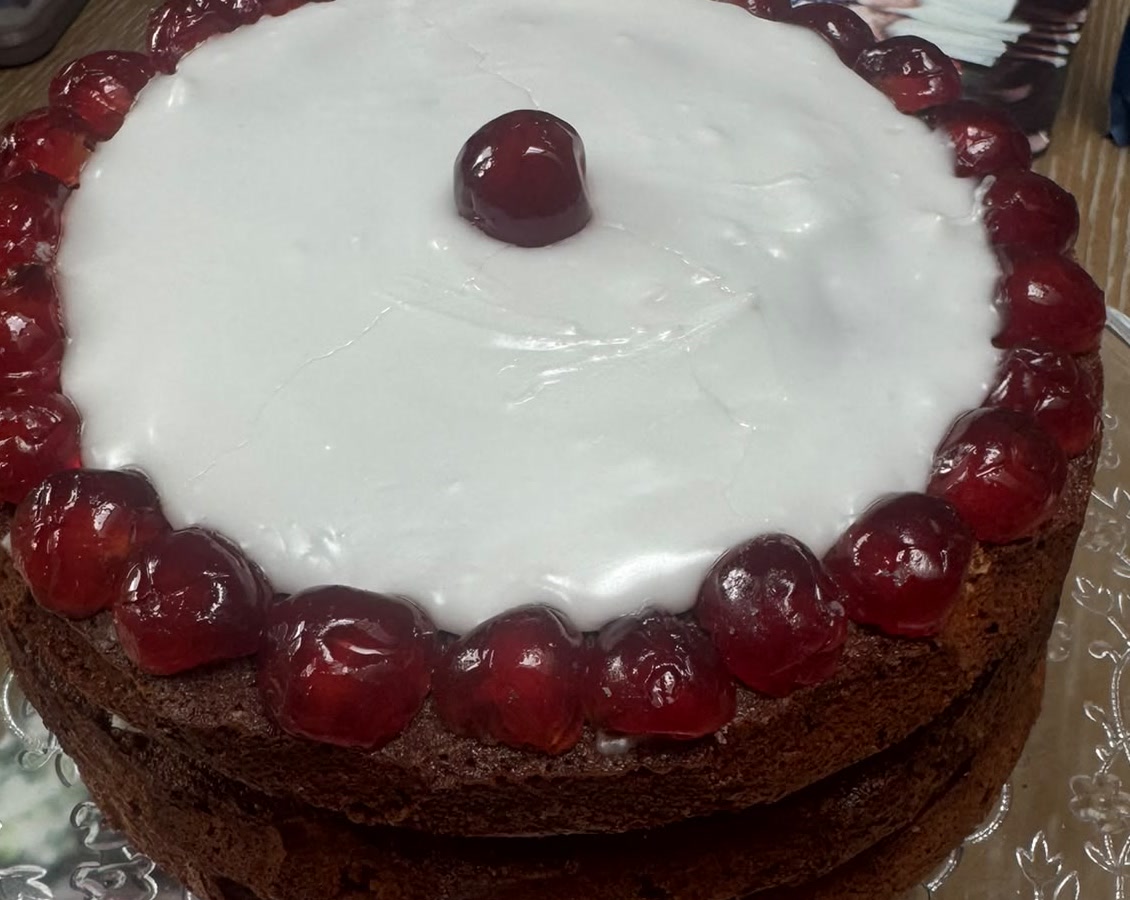 A large round cherry Bakewell cake on a glass cake stand, iced white with a ring of glace cherries around the edge and one in the middle. A large round cherry Bakewell cake on a glass cake stand, iced white with a ring of glace cherries around the edge and one in the middle.
