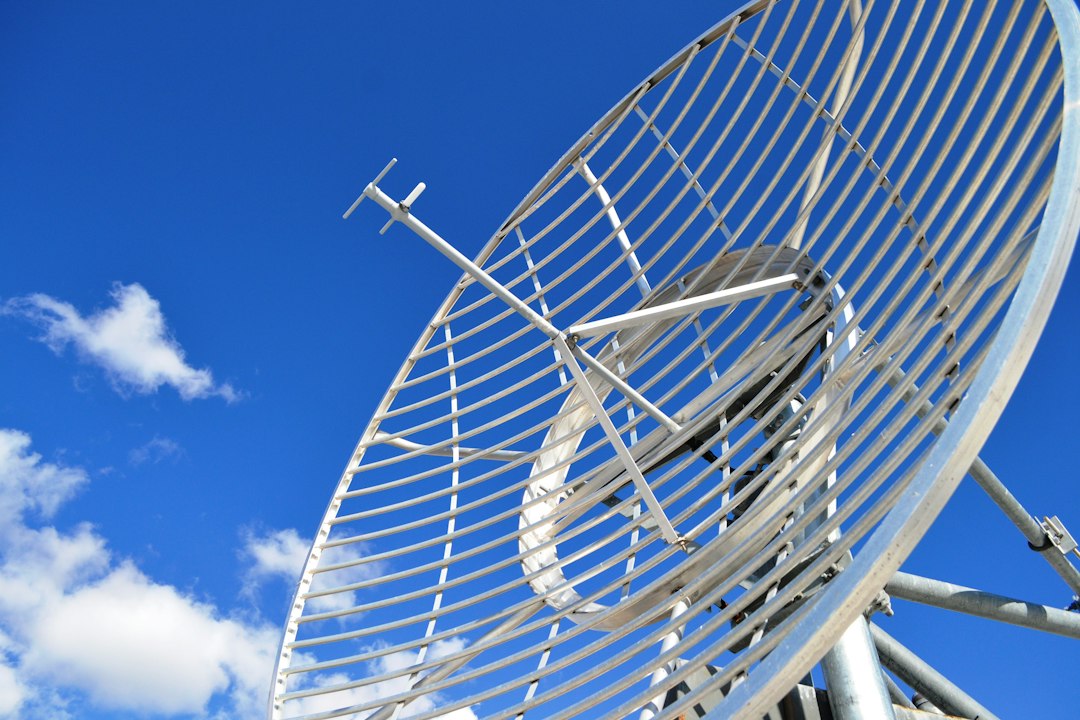 Low-angle view of a galvanized parabolic grid satellite antenna against a clear blue sky with a few clouds.