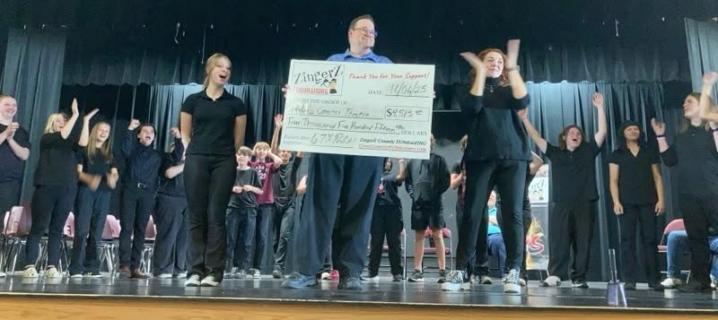 A cast and crowd on stage cheering as a big fundraising check is revealed