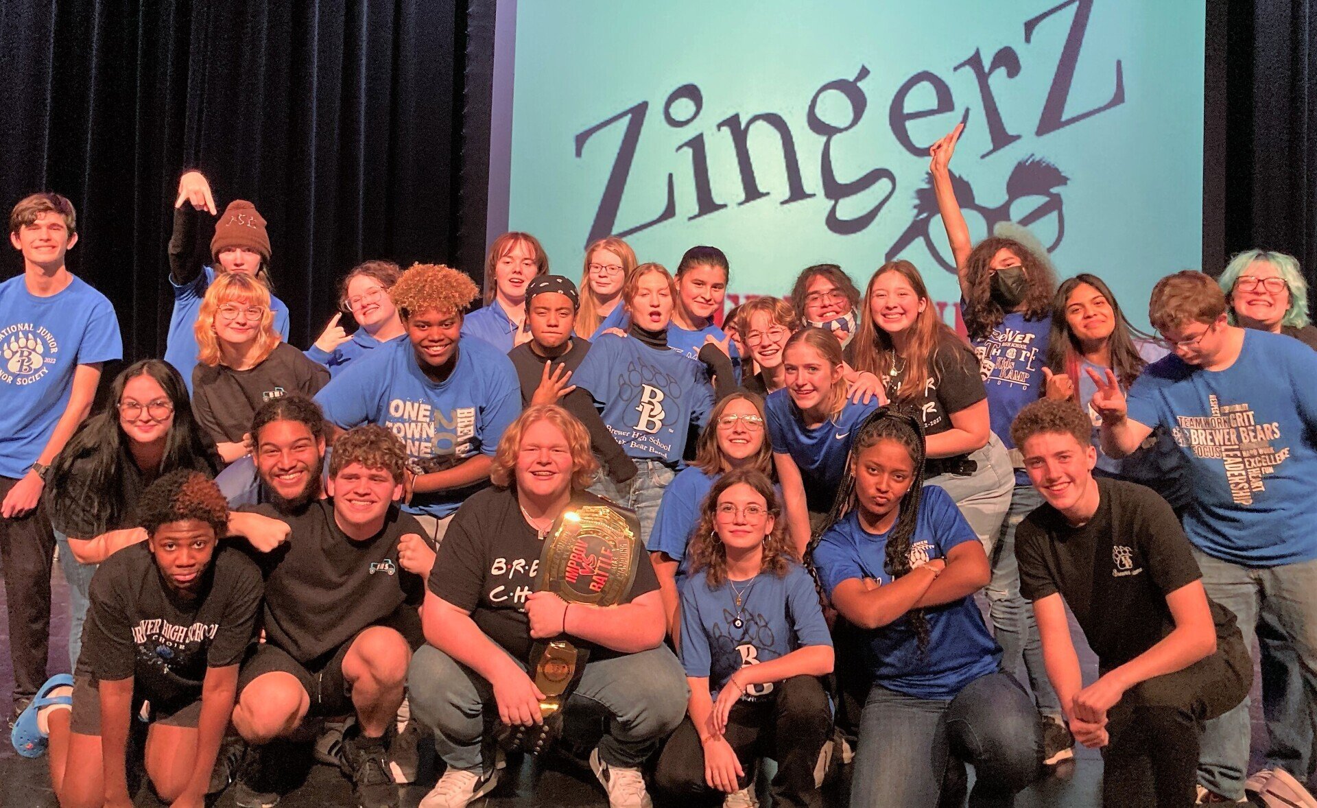 A high school cast crowded on stage holding the Improv Battle championship belt