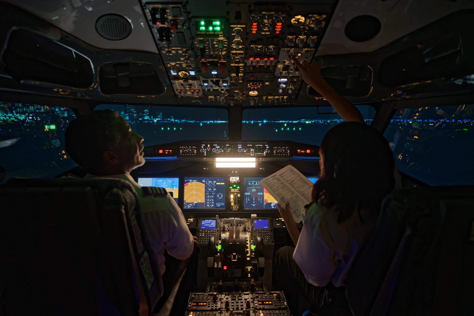 Two pilots on a flight deck at night, one reaching for the overhead panel