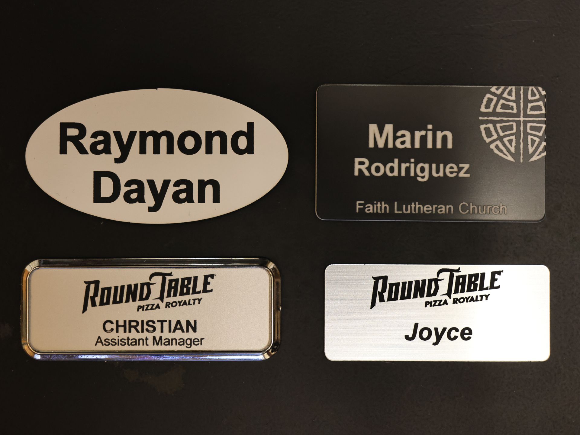 Four engraved name tags on a dark background in oval and rectangular formats, one in a polished metal frame, each carrying a name and logo