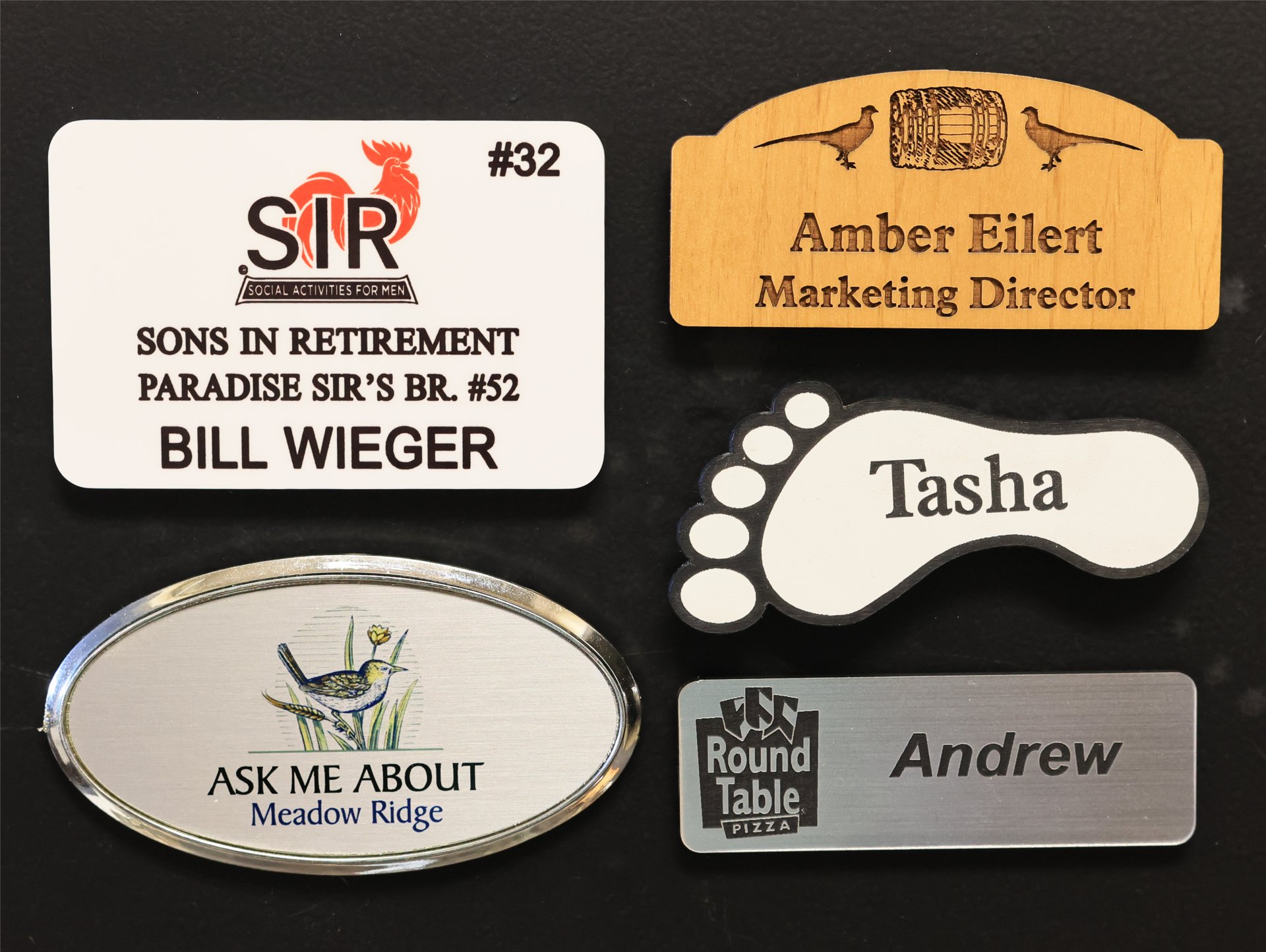 Six name tags in varied shapes and materials on a dark background, including plastic, wood, a shaped novelty badge and a framed oval
