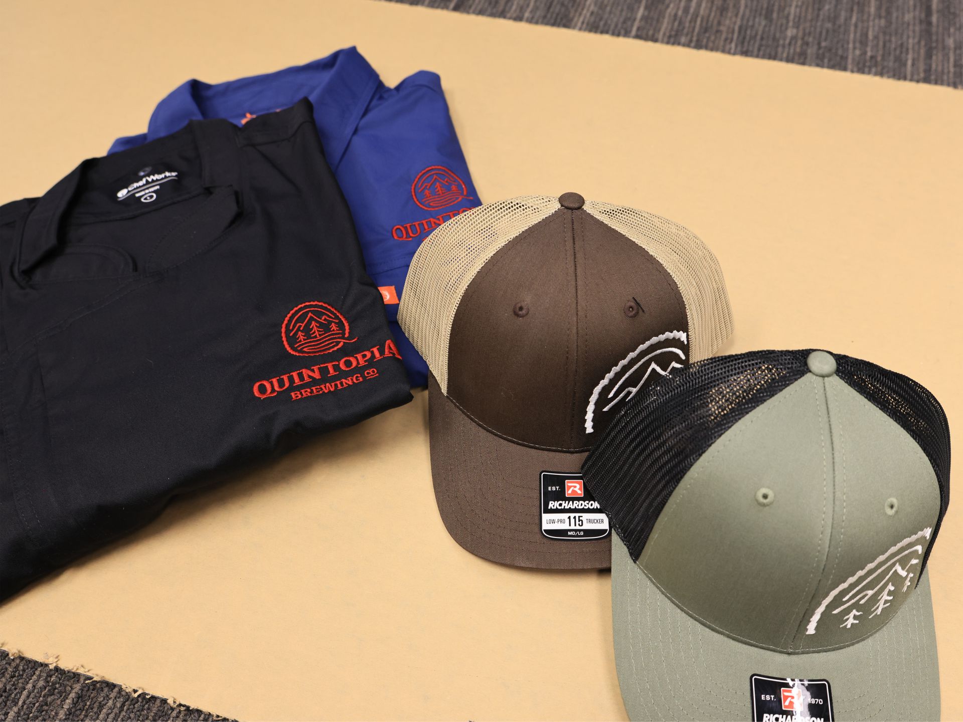Folded embroidered work shirts with a chest logo beside two embroidered trucker caps on kraft paper