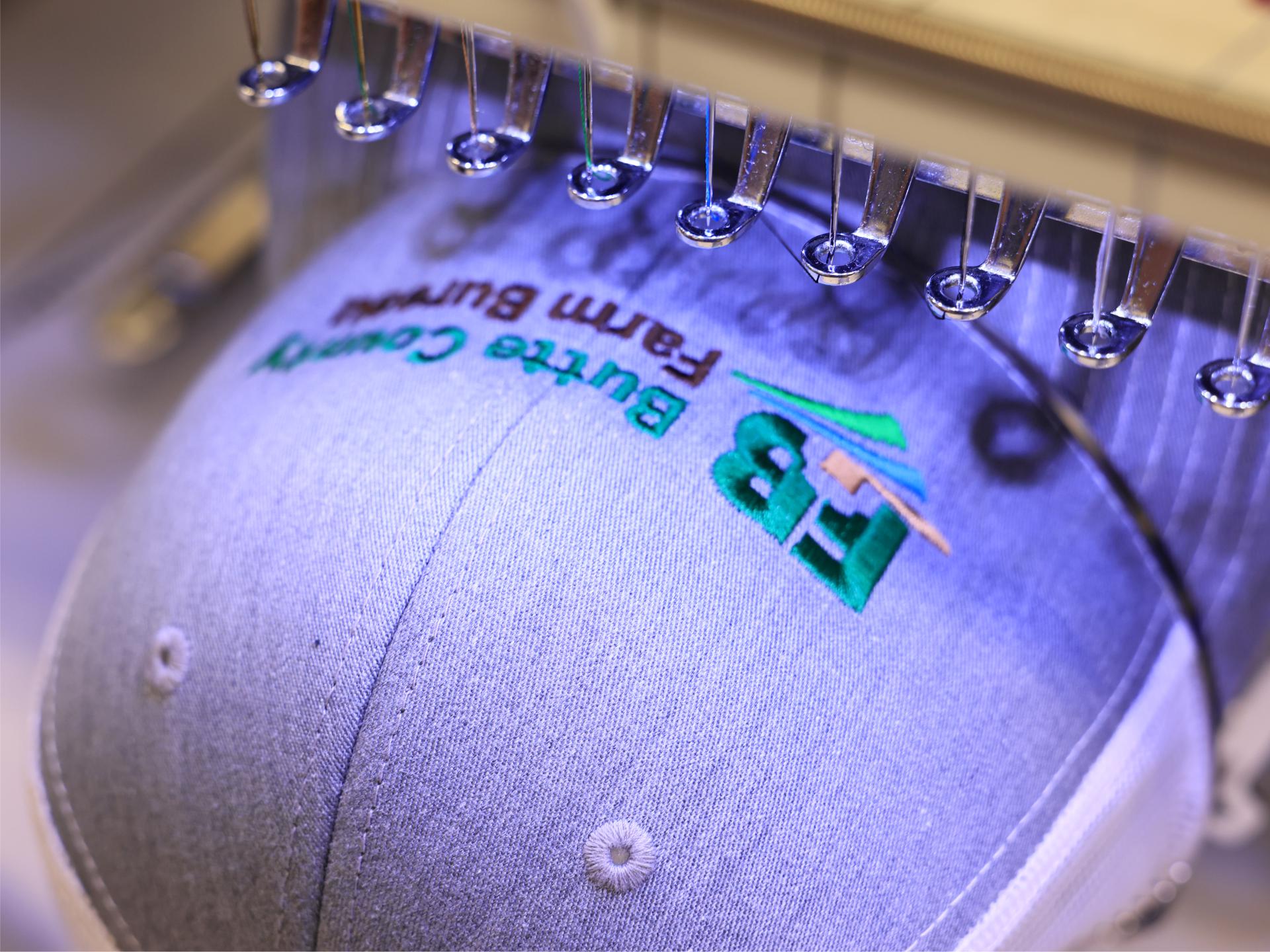 A cap held in the embroidery machine with its logo part stitched, needles above it