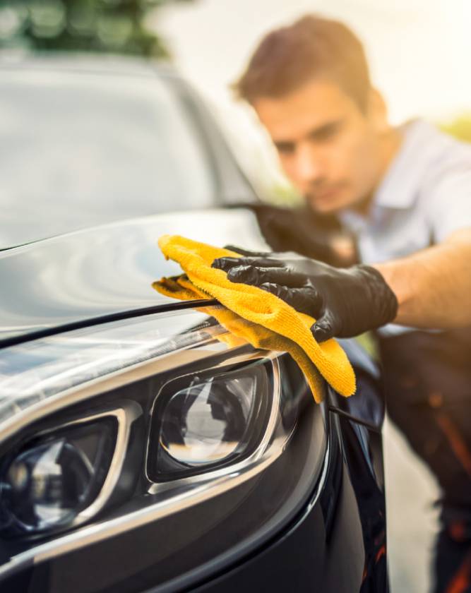 Technician in gloves wiping down the hood of a black car with a microfibre cloth after repair
