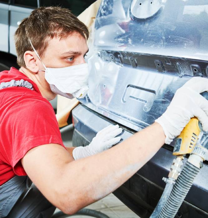 Technician in a dust mask and gloves block-sanding a body panel down to primer with a dust-extraction sander before refinishing