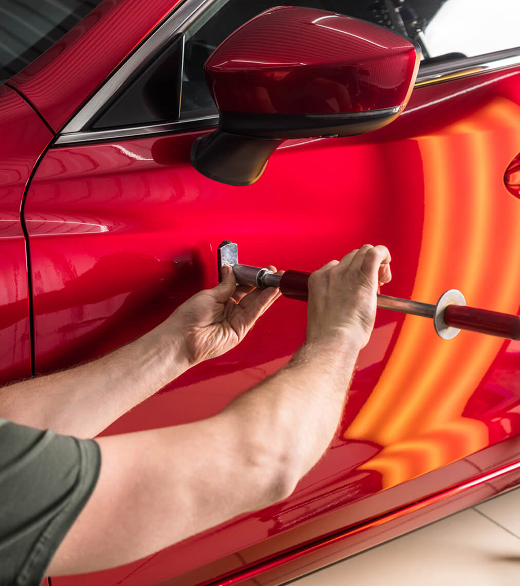 Technician using a paintless dent removal rod behind a red door panel, with a striped reflection board showing the contour of the metal
