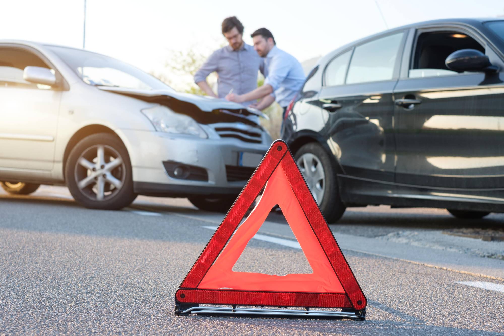 Two drivers inspecting a fender-bender between two cars, with a red emergency warning triangle set out on the road