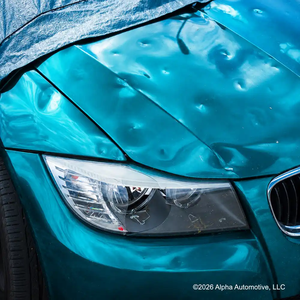 Hood and fender of a teal car covered in dozens of shallow round hail dents, with the paint finish unbroken