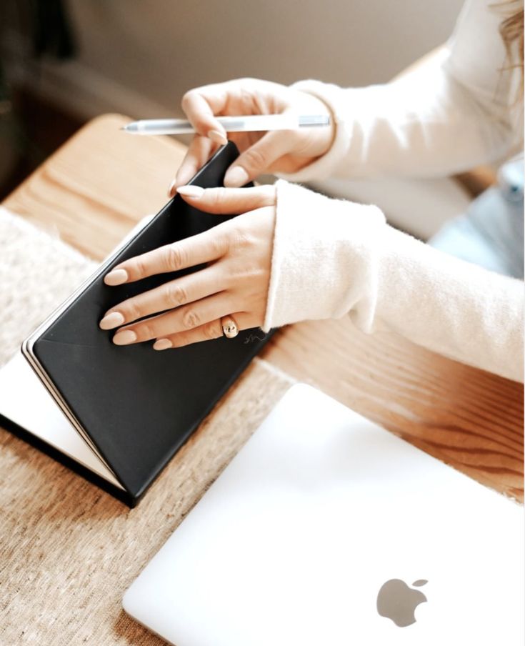 Une femme feuillette son calepn noir, un crayon à la main, avec un Macbook sur son bureau