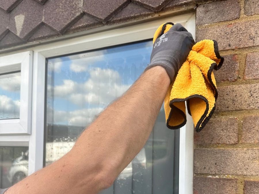 A gloved hand wiping a white window frame with a yellow microfibre cloth