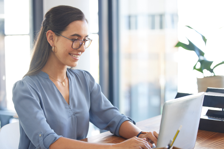 A woman wearing glasses and a blue shirt works on a laptop at a wooden desk.