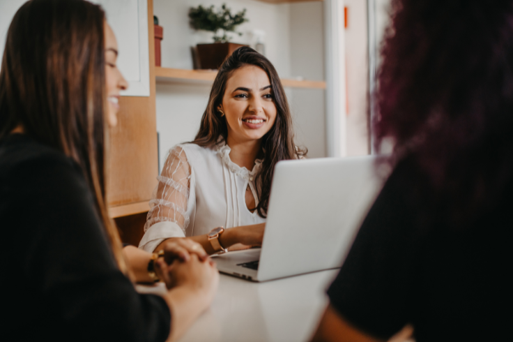 A woman in a white blouse smiles while looking at a laptop, with two other people sitting at the table.