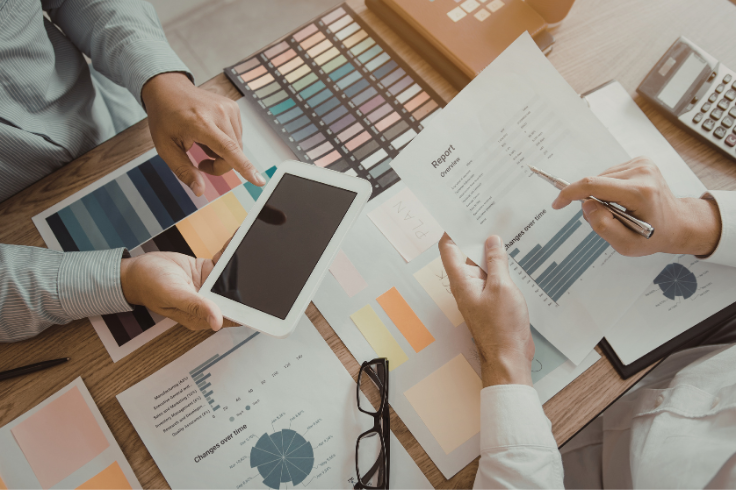 Hands pointing at a tablet, color swatches, reports, and a calculator on a wooden desk during a meeting. Hands pointing at a tablet, color swatches, reports, and a calculator on a wooden desk during a meeting.