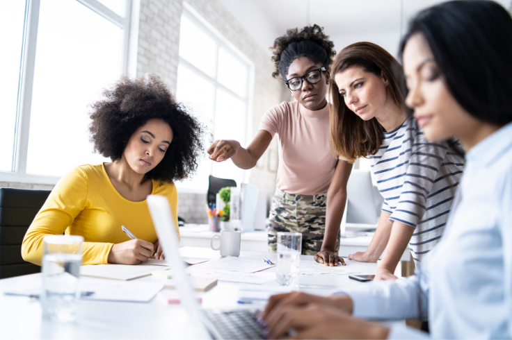 Four people collaborating around a table with laptops and papers, with one person writing and another pointing at something on the table. Four people collaborating around a table with laptops and papers, with one person writing and another pointing at something on the table.