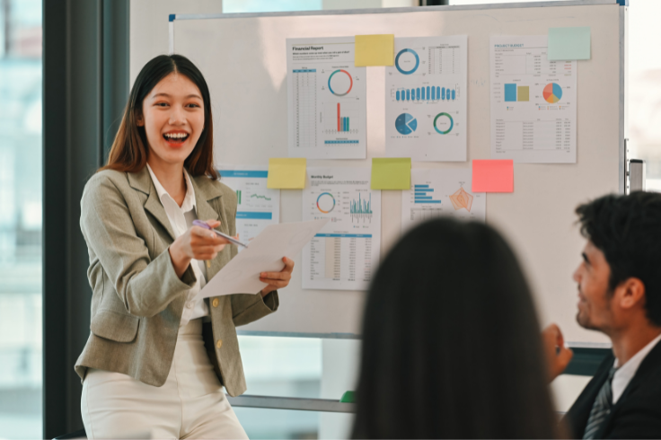 A woman in a suit smiles and gestures with a pen while holding papers in front of a whiteboard covered in charts and sticky notes. A woman in a suit smiles and gestures with a pen while holding papers in front of a whiteboard covered in charts and sticky notes.