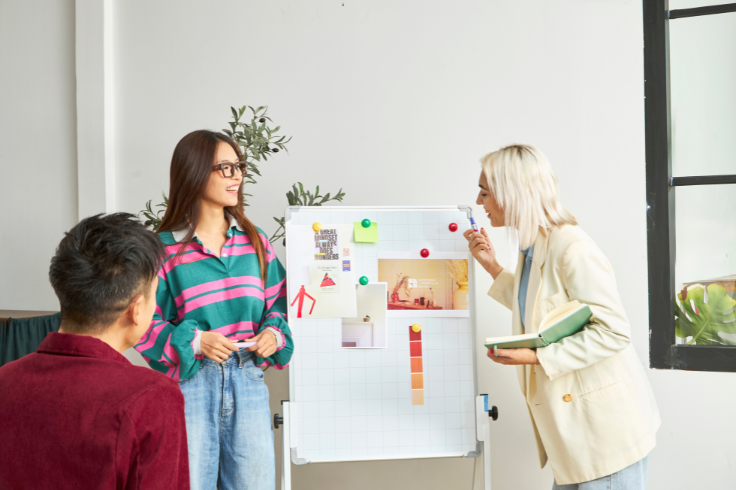 Three people are gathered around a whiteboard covered in notes and images, discussing ideas in a bright, modern office space. Three people are gathered around a whiteboard covered in notes and images, discussing ideas in a bright, modern office space.