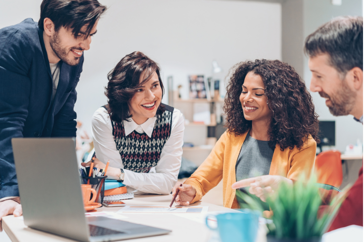Four colleagues are gathered around a table in an office, looking at papers and a laptop, and smiling. Four colleagues are gathered around a table in an office, looking at papers and a laptop, and smiling.