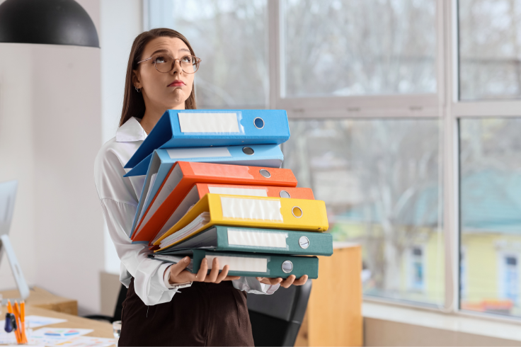 A person wearing glasses and a white shirt holds a tall stack of colorful ring binders, looking upwards with a frustrated expression. A person wearing glasses and a white shirt holds a tall stack of colorful ring binders, looking upwards with a frustrated expression.