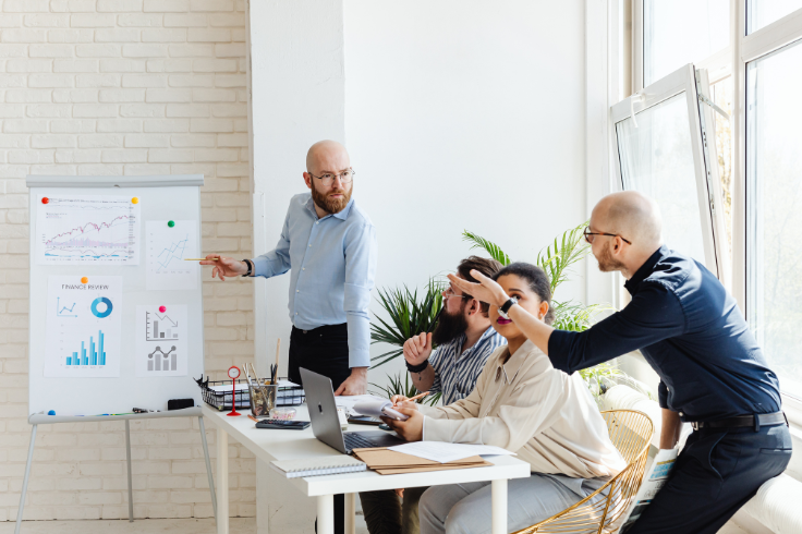 A man in a blue shirt points to a whiteboard with charts as colleagues listen at a table with laptops. A man in a blue shirt points to a whiteboard with charts as colleagues listen at a table with laptops.