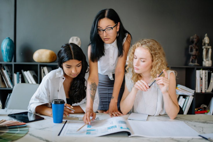 Three people gathered around a table, looking at documents and discussing them. One person is pointing at the papers. Three people gathered around a table, looking at documents and discussing them. One person is pointing at the papers.
