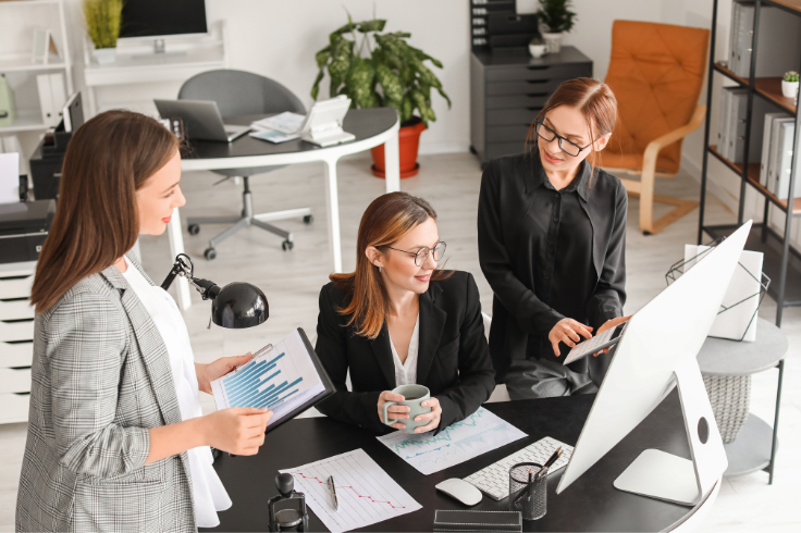 Three people in an office environment, two are looking at a computer screen, and one is holding a tablet, with charts and papers on the desk. Three people in an office environment, two are looking at a computer screen, and one is holding a tablet, with charts and papers on the desk.