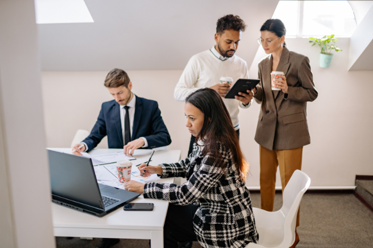 Four people in business attire gathered around a table in an office setting, working on documents and a tablet. 