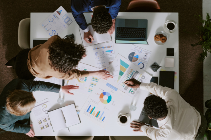 A group of people are gathered around a table, looking at charts and graphs scattered across the surface, with a laptop and coffee cups visible. 