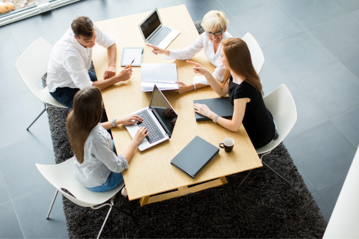 A group of people are gathered around a wooden table in a modern office space, engaged in a discussion with laptops and documents spread out before them.