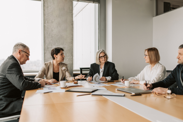 A group of people in business attire sit around a conference table, reviewing documents and engaging in discussion. 