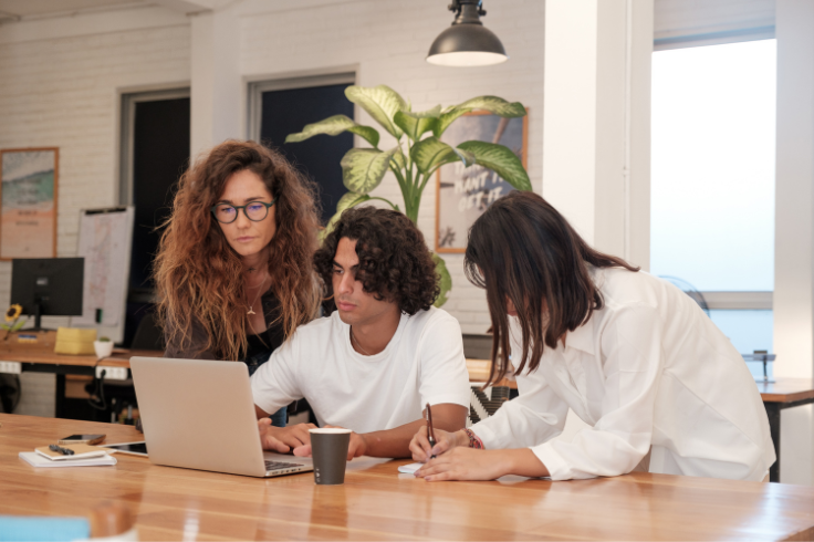 Three people are gathered around a laptop on a wooden table in an office setting.