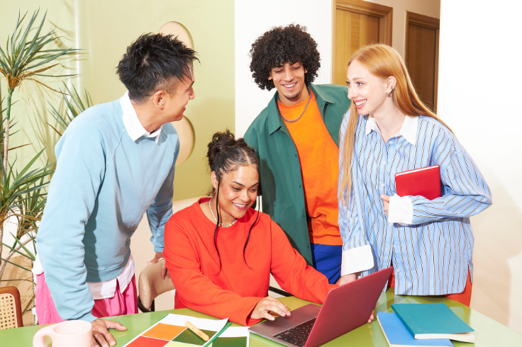 Four people gathered around a table, looking at a laptop. One person is typing on the laptop, while the others look on and smile. 