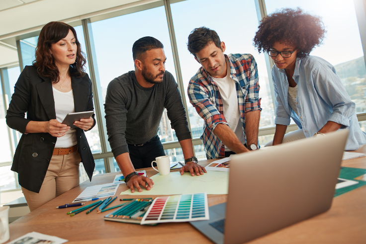 A diverse group of people collaborating around a table with a laptop, tablet, and color swatches. 