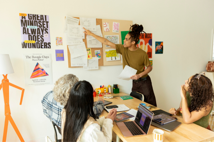 A person stands and points to a corkboard covered in papers and colorful artwork while others sit at a table with laptops and tablets. 